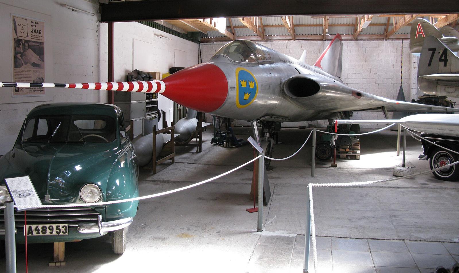 Grønn Saab 92 i en museumshall, ved siden av et sølvfarget jagerfly med rød nese