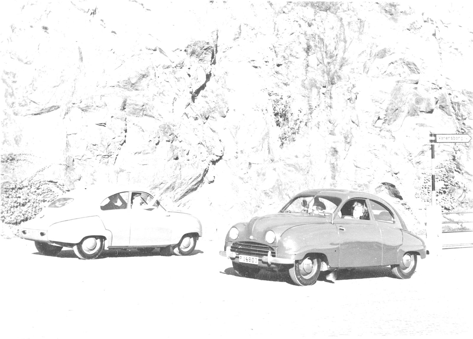 Svart-hvitt arkivbilde av to Saab 92 foran en bergvegg, med et veiskilt mot Vänersborg
