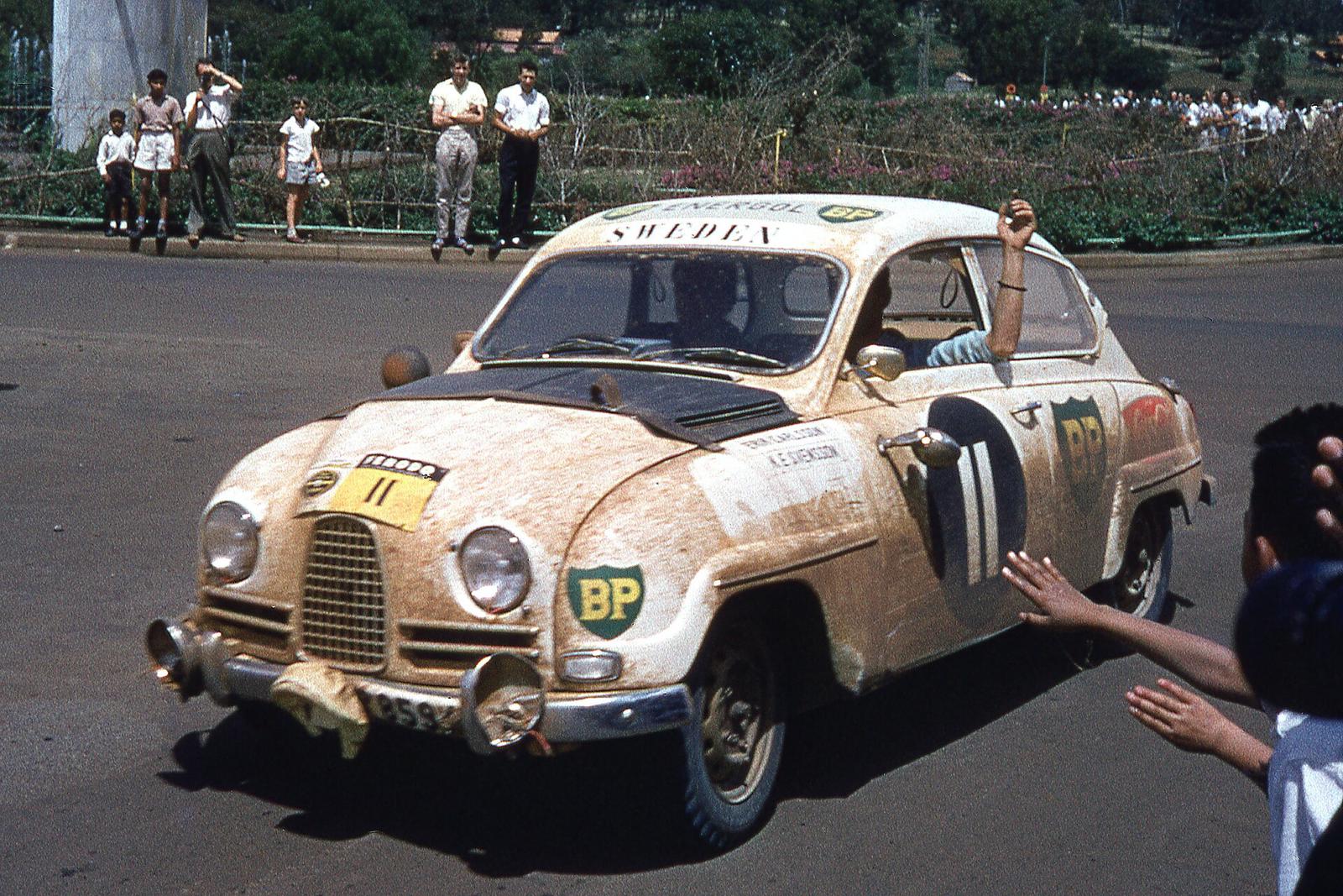 Skitten, lys Saab 96 med startnummer 11 og BP-merker kjører forbi tilskuere på en gate