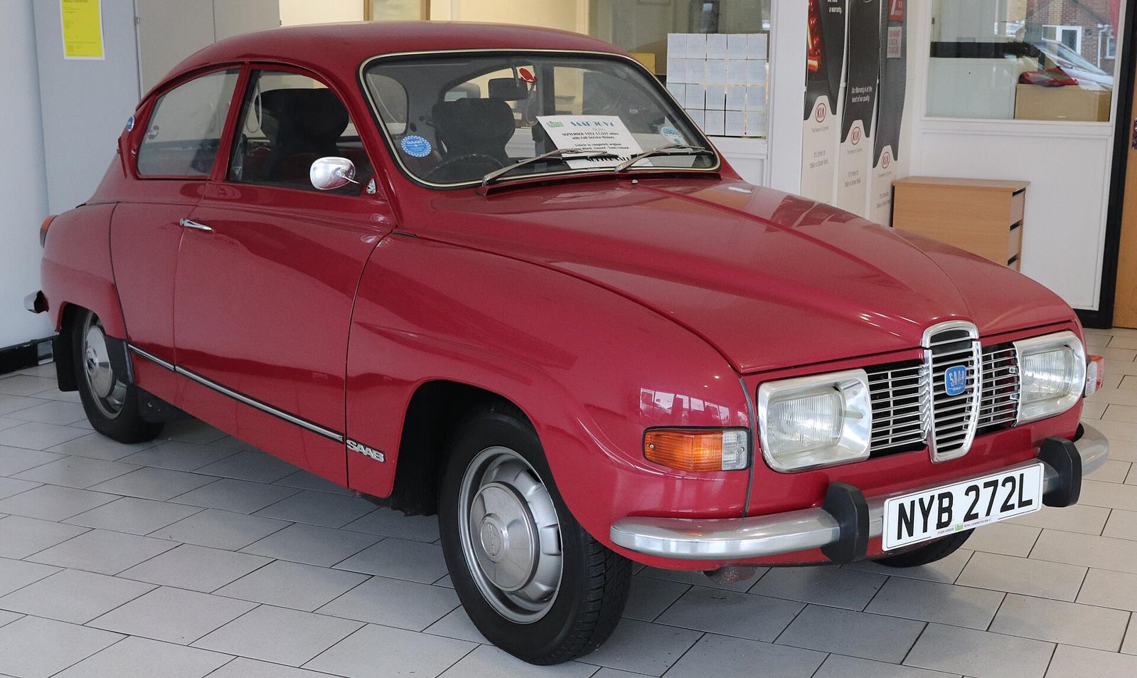Rød Saab 96 V4 med rektangulære lykter, sett skrått forfra i et utstillingslokale