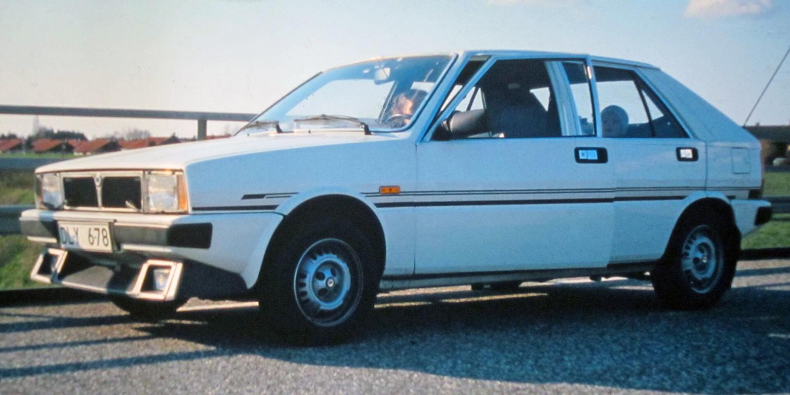 Hvit Saab-Lancia 600 sett skrått forfra, parkert ved et autovern med røde hustak i bakgrunnen