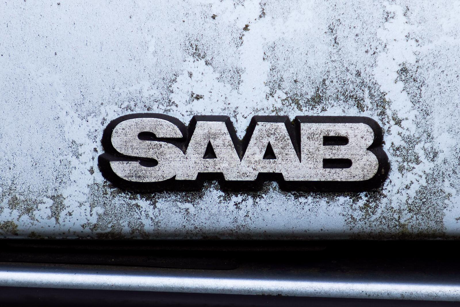 Værbitt SAAB-skriftmerke på siden av en hvit bil, med mose på lakken