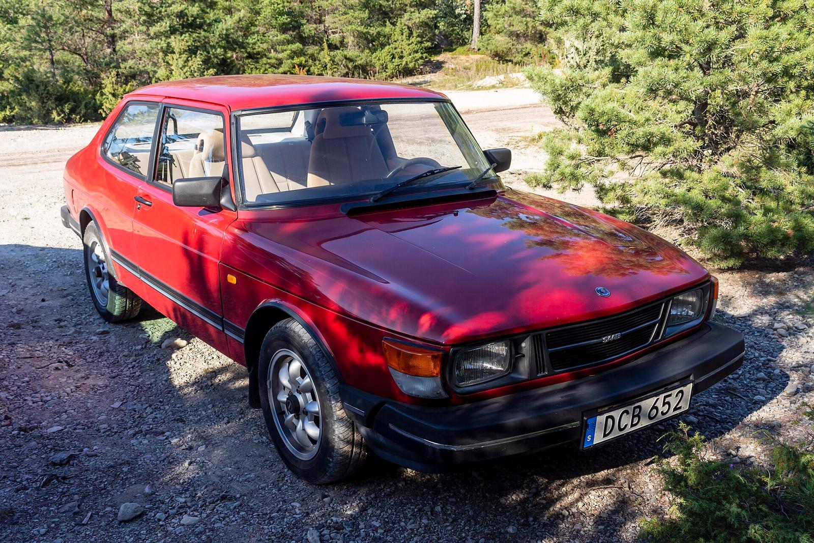 Rødbrun Saab 90 sett skrått forfra, parkert på grus foran furutrær