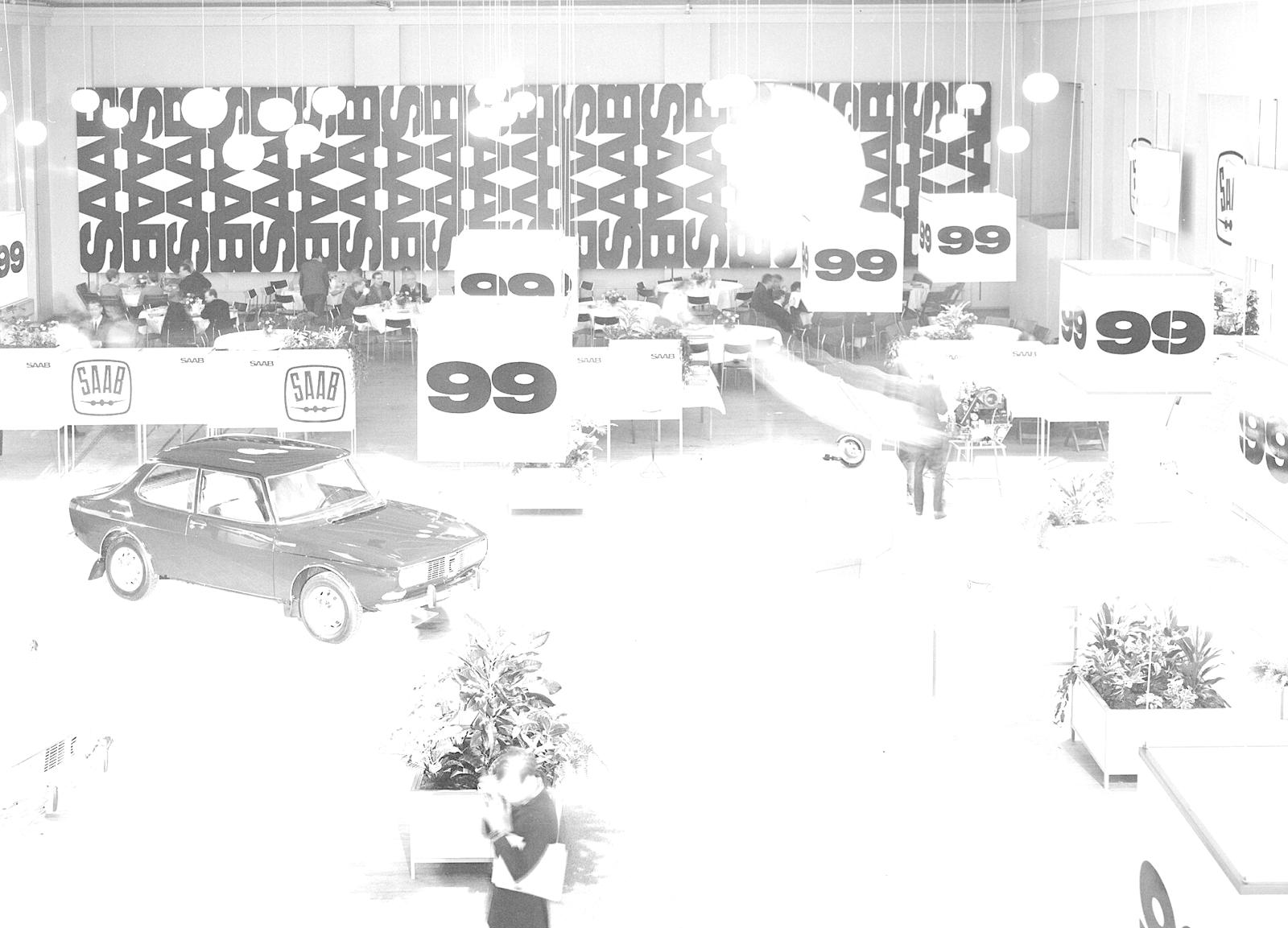 Svart-hvitt bilde fra en utstillingshall med en Saab 99 på gulvet og store skilt med tallet 99