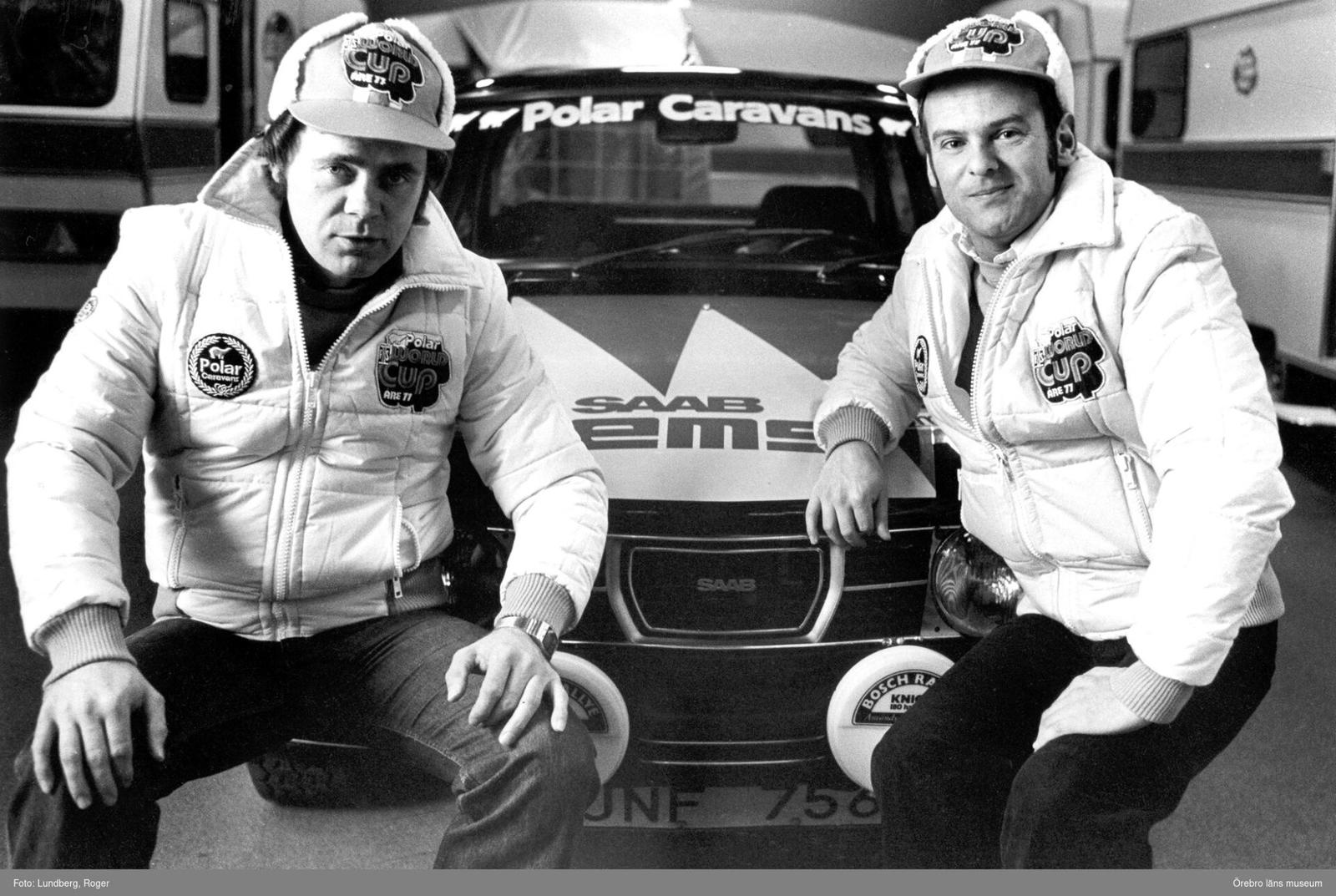 Svart-hvitt bilde av to rallyførere i hvite jakker foran fronten på en Saab 99 EMS rallybil